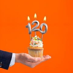Hand holding birthday cupcake with number 120 candle - background orange