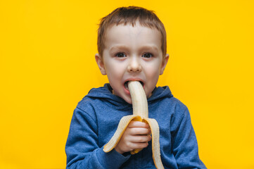 A small funny cheerful boy bites a banana, makes faces and plays around. Studio photography with yellow background.