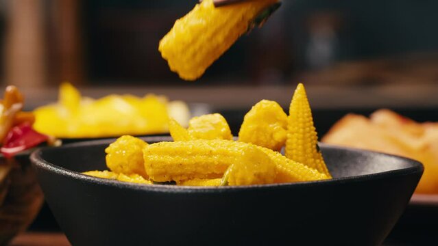 Close-up of fermented yellow mini corn with black pepper on black plate. Preservation of vegetables in glass jars. Fermentation preserved mini corns with spices.