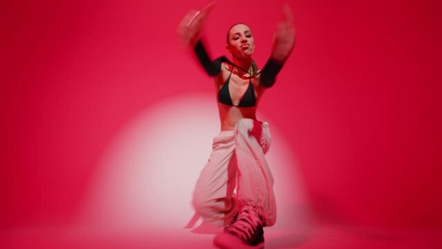 Brunette female dancer dancing moves energetically performing hip hop dance on red color wall round white spot light background in studio. Sexy woman demonstrates modern contemporary choreography