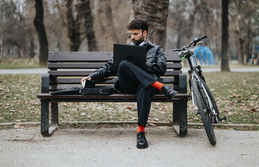 Elegant male business entrepreneur working remotely on his laptop in a city park, with a bicycle beside him on the bench.