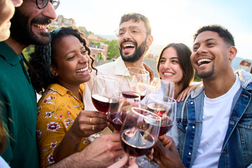 Cheerful groups of international friends toasting glasses of red wine together laughing at terrace bar outdoors. Close up cheers of smiling diverse people having fun at sunset party. Summer vibes
