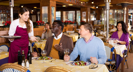 Positive waitress and two man of the restaurant pay for dinner using smartphones