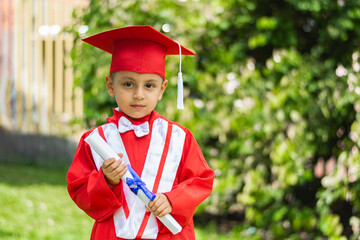portrait of little latin boy in red graduation gown, posing while holding diploma, in the park