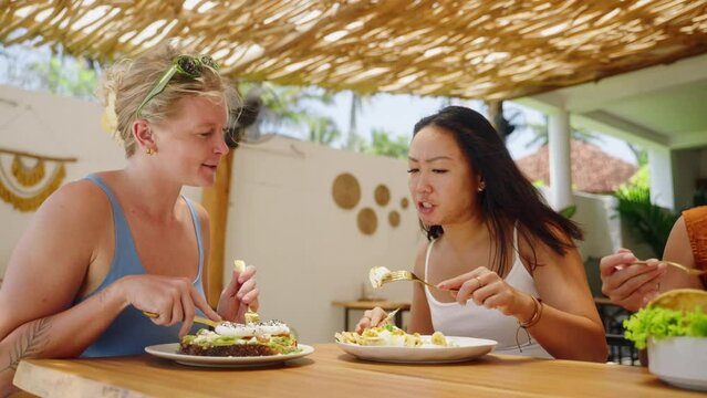 Two pretty women eating, talking about food in hipster cafe on Bali. Diverse female friends enjoying brunch in tropical open air restaurant. Caucasian, asian girls having lunch on the weekend.