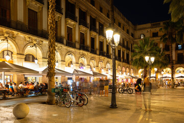 Evening view of bustling Placa Reial in Barcelona with well-lit cafes and people enjoying leisurely...