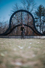 A casually dressed young man rides his bicycle across a unique, bridge-like structure in the park during a bright, sunny day.