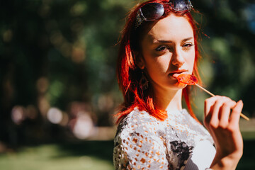 A gorgeous young redhead girl posing with a lollipop in a sunny park, surrounded by lush greenery, embodying a carefree summer vibe.