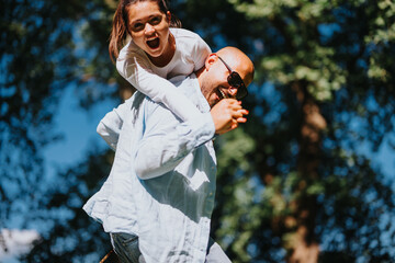 A cheerful moment as a father with sunglasses gives a piggyback ride to his delighted daughter in an outdoor sunlit park.