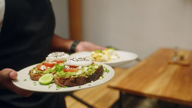 Waiter hands carry vegan food, avocado toasts on plates for happy young women in cafe. Waiter serving healthy food to clients. Diverse female friends eat tasty dishes in the restaurant. Close-up.