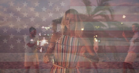 Fototapeta premium Image of flag of united states of america over happy diverse friends with sparklers on beach