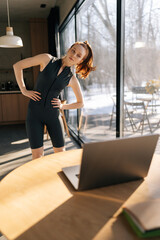 Vertical shot of sporty young woman working out, doing warming up exercise watching fitness video online on laptop at home standing by window on sunny day. Female practicing exercise with laptop