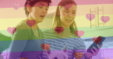 Image of heart emojis and rainbow flag over caucasian female couple using smartphone