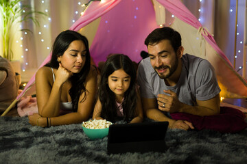 Happy family enjoying a movie night together in a homemade tent with popcorn and a tablet, creating...