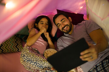 Father and daughter enjoying movie night under a cozy blanket fort at home, using a tablet