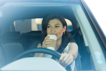 Confident young woman sipping coffee and smiling while driving in her car, captured through the...