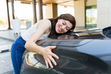 Happy woman lovingly hugs the hood of her shiny car, expressing affection and contentment with her...