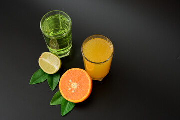 Two tall glasses with different citrus juices on a black background, next to the halves of ripe orange and lime with leaves.