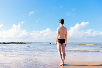 Man wearing swimwear standing on the sand of a beach. He is standing looking at the sea and the...