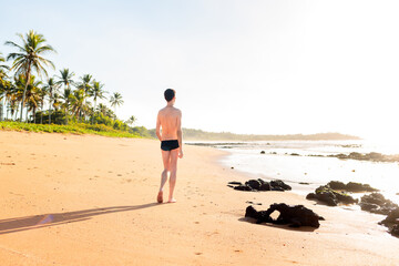 Man wearing swimwear walking calmly on the sand of a paradisiacal beach, with coconut trees and sunrise in the background.