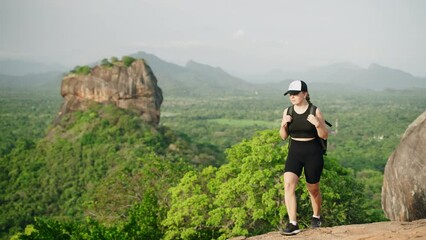 Active female hiker in sportswear climbs mountain trail near Sigiriya Rock. Adventurous traveler with backpack enjoys Sri Lanka scenery, explores ancient landmarks, experiences solo journey on foot.