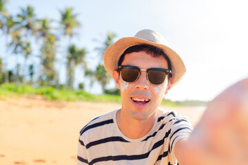 A man in beach wearing hat and sunglasses smiles, taking a selfie on a paradisiacal beach, capturing the essence of summer vacation joy. He is using sunscreen on his face.