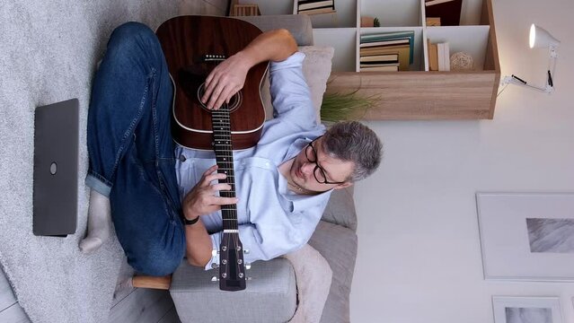 Vertical video. Home education. Musician student. Online lesson. Smart concentrated man practicing acoustic guitar chords sitting at laptop meeting on living room floor copy space.
