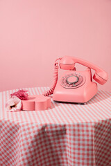 Pink color concept with a table covered by pink checkered tablecloth containing a desk phone next to an empty pedestal in flower shaped. Photo was taken from front view, copy space