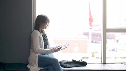 Woman sitting by a large window reading a book, basking in natural light. She wears casual attire and is seated comfortably, absorbed in her reading
