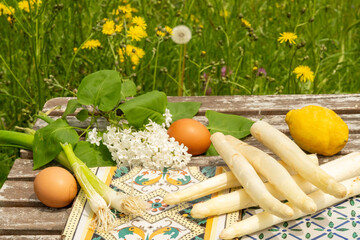 Seasonal cuisine: fresh asparagus stacked on colorful tiles. Together with eggs, Spring onions, a lemon and lilac blossom on a wooden table which is placed in the garden