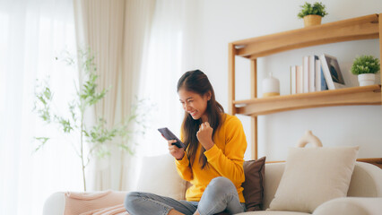 Happy young asian woman relax on comfortable couch at home texting messaging on smartphone, smiling...