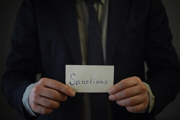 Man showing sanctions word on paper and devastated middle aged caucasian man in a black suit composition with dramatic lighting