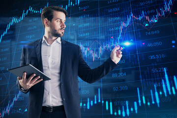 Attractive young european businessman with tablet using creative candlestick forex chart with index on blurry office background. Trade and stock exchange concept. Double exposure.