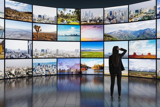 Back view of young businesswoman looking at video wall in television production room as technology concept with colorful screens and various images. Monitors on light rooms with reflections on floor.