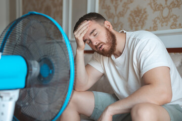Young bearded man using electric fan at home, sitting on couch cooling off during hot weather,...