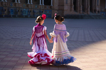 Two pretty little girls dancing flamenco dressed in typical gypsy costumes walk hand in hand through a famous square in Seville. The girls look at each other happily. Photo taken from behind.
