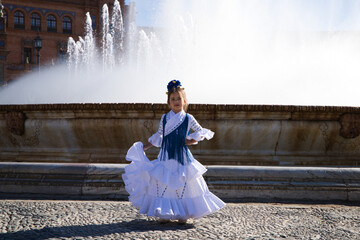 A pretty little girl dancing flamenco dressed in a white dress with ruffles and blue fringes in a...