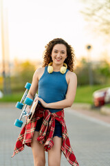 Cheerful young woman with curly hair, holding a skateboard and wearing headphones, smiling in a...