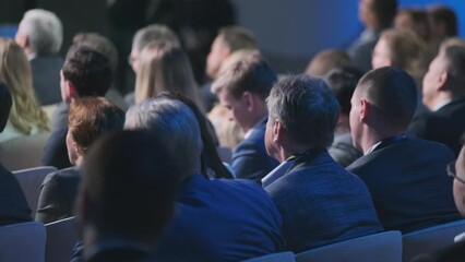 Back view of diverse participants focused and listening at a business conference. The setting suggests a professional environment, engaging with the speaker on stage.