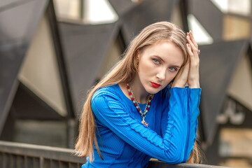 Fototapeta premium An elegant girl poses in a blue dress on a bridge between buildings on the street.