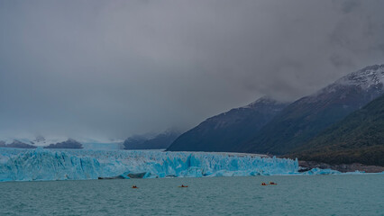 A wall of cracked blue ice. The glacier stretches to the horizon between the mountains. Canoes with...