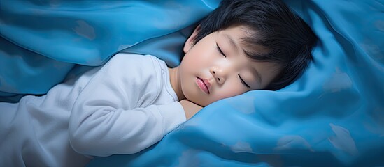A charming young Asian toddler aged 3 4 peacefully rests on a blue pillow in bed wearing pajamas This delightful image embodies the concept of bedtime for children and offers space for copy