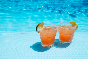 Two glasses with a tasty cocktail with ice cubes. Delicious fresh drink with lime slices by the pool. A sunny day and the reflection of rays in the blue water in the background.
