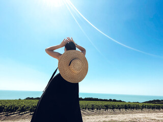 Young woman in black long dress and wide brimmed straw hat stands in graceful pose and looks at...