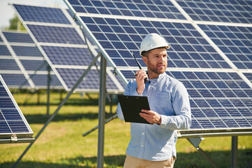 Using walkie talkie and holding notepad. Engineer with photovoltaic solar panels outdoors at daytime