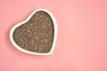 Photograph of chia seeds in heart shape white bowl with copy space on pink colour background. Top view. Health care concept.