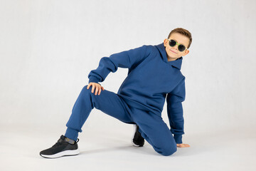 Cheerful boy in a blue tracksuit and sunglasses. Pretty boy,. Baby model. A little boy poses on a white background. Boy posing sitting.Dancing boy