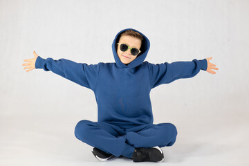 Cheerful boy in a blue tracksuit and sunglasses. Pretty boy,. Baby model. A little boy poses on a white background. Boy posing sitting