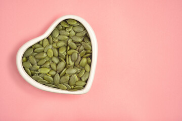 Photograph of  hulled green pumpkin seeds in heart shape white bowl with copy space on pink colour...