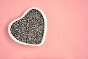 Photograph of poppy seeds in heart shape white bowl with copy space on pink colour background. Top...